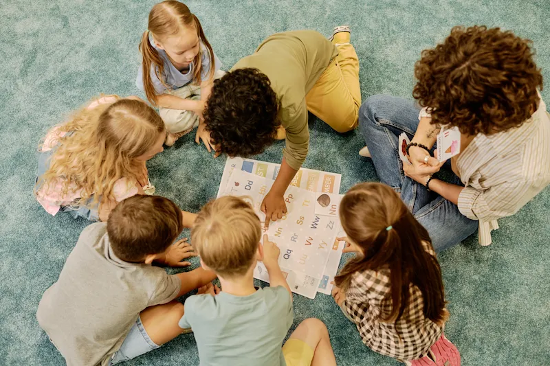 professor e alunos sentados no chao observadno livro de ingles
