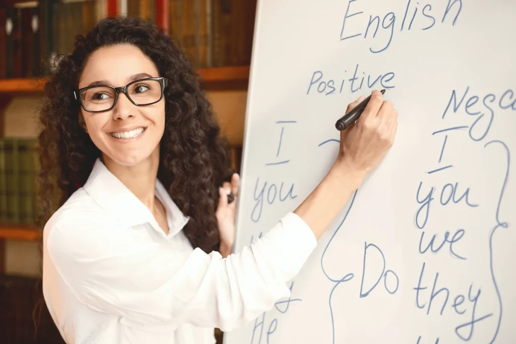 professora de ingles escrevendo palavras no quadro e sorrindo para a turma